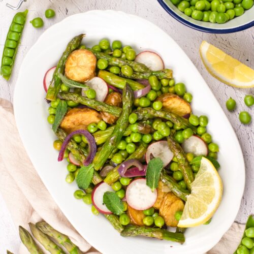 Plate of roasted asparagus, peas, radishes, and lemon wedges on a white platter with mint garnish, ready for serving